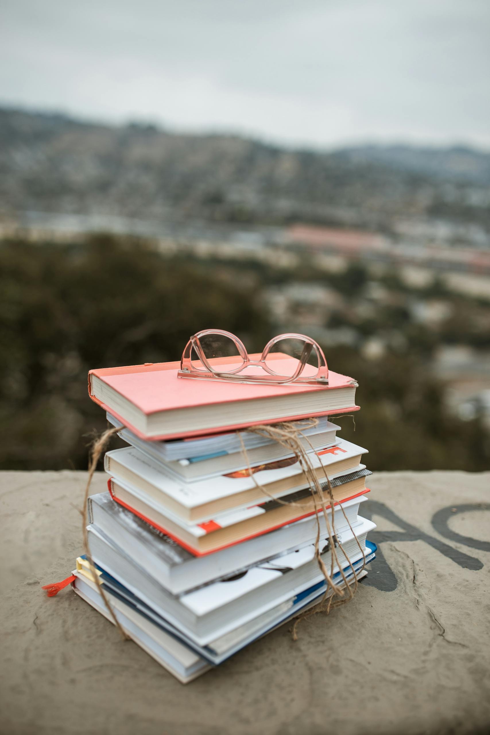 Stack of colorful books with glasses on top, set outdoors with blurred background.